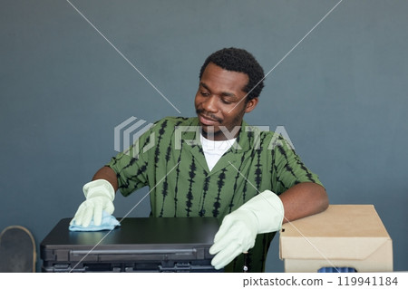 Waist up portrait of smiling African American man cleaning office equipment and wearing gloves on simple blue background, copy space 119941184
