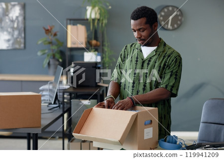 Waist up portrait of young African American man unpacking boxes on table in empty office, copy space 119941209