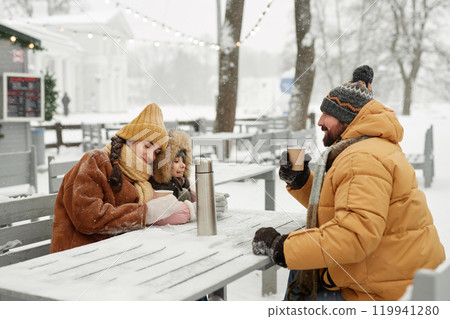 Portrait of modern young family enjoying hot drinks sitting at outdoor cafe table with thermos enjoying winter activities together 119941280