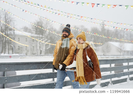 Waist up portrait of happy young couple holding hands walking by skating rink in winter park and enjoying outdoor activities with snow falling copy space 119941666