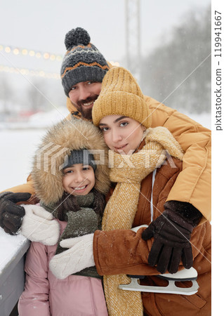 Vertical waist up portrait of modern young family with daughter posing outdoors in winter park together and looking at camera smiling happily 119941667