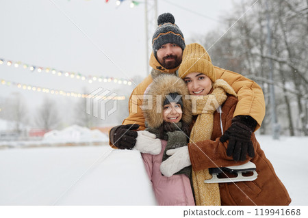 Waist up portrait of happy young family with daughter standing by skating rink in winter park together and looking at camera smiling copy space Waist up portrait of happy young family with daughter standing by skating rink in winter park together and looking at camera smiling copy space 119941668