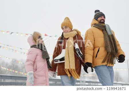 Low angle portrait of smiling young family with child holding hands and walking towards skating rink in snowy winter park 119941671