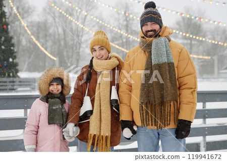 Front view portrait of happy young family looking at camera and smiling while standing by skating rink in winter park on Christmas 119941672