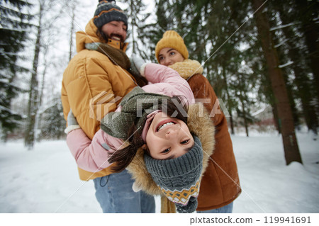 Portrait of happy young family playing with daughter in winter forest focus on cute girl hanging upside down in fathers arms and smiling at camera Portrait of happy young family playing with daughter in winter forest focus on cute girl hanging upside down in fathers arms and smiling at camera 119941691