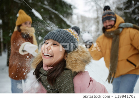 Close up action shot of smiling young girl playing snowball fight with family in winter forest and running away Close up action shot of smiling young girl playing snowball fight with family in winter forest and running away 119941702