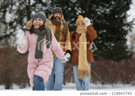 Candid shot of cute young girl running in snowy forest with parents in background happy family enjoying holidays outdoors copy space 119941743