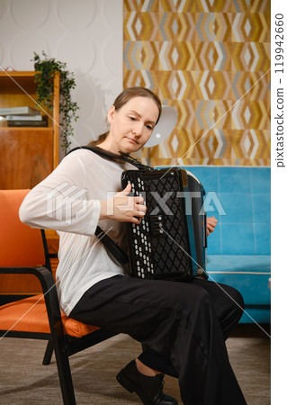 A middle-aged woman playing the accordion while seated in a modern living room with colorful decor A middle-aged woman playing the accordion while seated in a modern living room with colorful decor 119942660