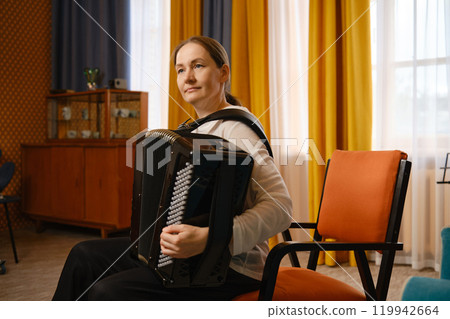 A middle-aged woman plays the accordion in a cozy room with colorful curtains and soft lighting 119942664