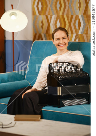Middle-aged woman enjoys playing her accordion while sitting on a stylish blue couch at home 119942677