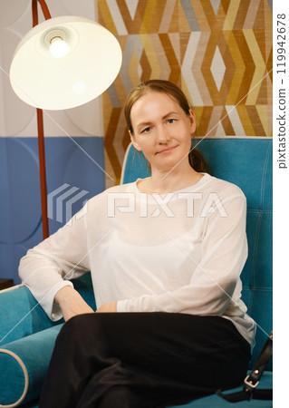 Casual portrait of a middle-aged woman relaxing in a blue armchair in a stylish interior  119942678
