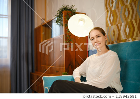 A middle-aged woman relaxes on a cozy couch in a stylish room with modern decor and warm lighting 119942681