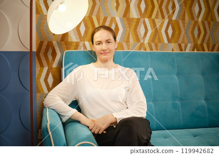 A middle-aged woman sits elegantly on a turquoise couch with geometric wallpaper in a modern living space 119942682