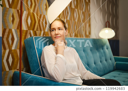 A thoughtful middle-aged woman seated on a stylish blue sofa in a cozy, modern living room setting 119942683