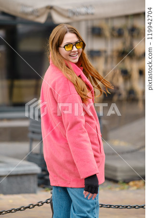 Young woman in bright pink jacket and sunglasses poses happily outdoors on a sunny day 119942704