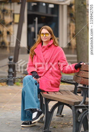Woman in a pink coat and yellow sunglasses sitting on a bench in an urban park during fall Woman in a pink coat and yellow sunglasses sitting on a bench in an urban park during fall 119942705