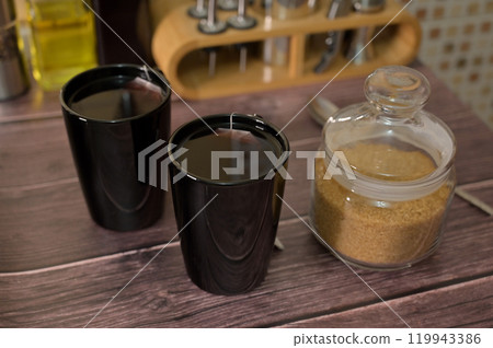 Two black cups with hot tea on wooden table, brown sugar in a jar Two black cups with hot tea on wooden table, brown sugar in a jar 119943386