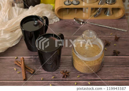 Two black cups with hot tea on wooden table, brown sugar in a jar, spicy spices and cinnamon 119943387
