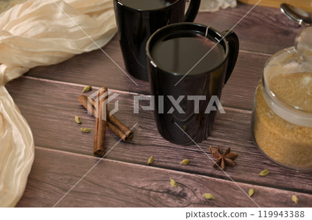 Close-up two black cups with hot tea on wooden table, brown sugar in a jar, spicy spices and cinnamon, cloth on background Close-up two black cups with hot tea on wooden table, brown sugar in a jar, spicy spices and cinnamon, cloth on background 119943388