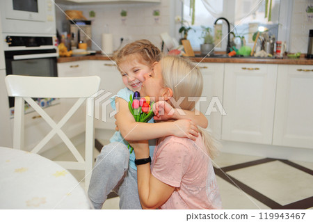 a joyful moment between a mother and her daughter as they embrace each other, holding vibrant flowers in their hands. Set against the backdrop of a bright, white kitchen, their smiles radiate love and 119943467