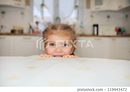 a young girl peeking out from behind a table in a bright, white kitchen, her eyes full of excitement and anticipation for a delicious meal.The clean kitchen backdrop enhances the feeling of a 119943472