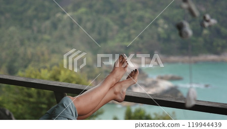 A person is seen relaxing with their feet propped up on a rail, enjoying a quiet moment in a serene setting. 119944439