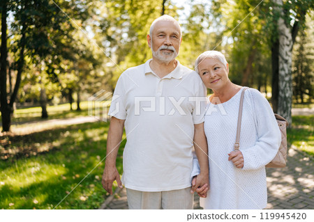 Portrait of loving senior couple enjoying golden years together, holding hands, posing standing in green summer park on sunny day, embodying togetherness and healthy aging with joy and contentment. 119945420