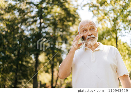 Low-angle view of elderly male looking peaceful and happy chatting on mobile phone in sunny summer park, harmonizing nature with technology in tranquil, joyful moment. Concept of happy retirement life Low-angle view of elderly male looking peaceful and happy chatting on mobile phone in sunny summer park, harmonizing nature with technology in tranquil, joyful moment. Concept of happy retirement life 119945429