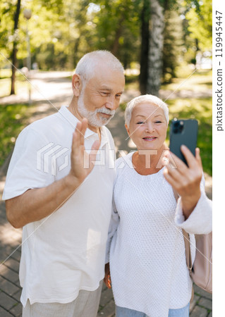 Vertical portrait of happy senior couple enjoying video call in summer sunny park, having smiling, laughing and cherishing moments in retirement, embracing technology and staying connected outdoors. Vertical portrait of happy senior couple enjoying video call in summer sunny park, having smiling, laughing and cherishing moments in retirement, embracing technology and staying connected outdoors. 119945447