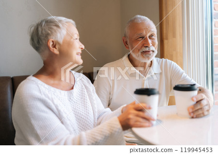 Loving senior couple enjoying cozy cafe date, smiling and chatting over coffee sitting hugging by window, showcasing enduring love and happiness. Concept of active aging and happy golden years. 119945453