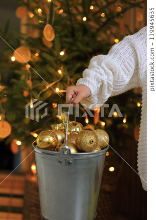 a pair of hands holding an array of shimmering golden ornaments, perfect for decorating a Christmas tree. The warm glow of twinkling lights and festive decorations in the background enhances the a pair of hands holding an array of shimmering golden ornaments, perfect for decorating a Christmas tree. The warm glow of twinkling lights and festive decorations in the background enhances the 119945635