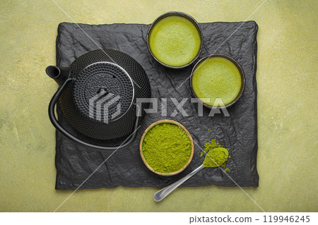 Matcha tea powder and hot drink in bowls with black iron teapot on black slate on green background, Japanese tea top view 119946245