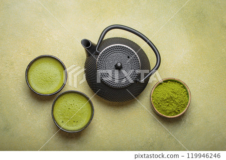 Matcha tea powder and hot drink in bowls with black iron teapot on green background, Japanese tea top view 119946246