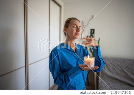 Relaxed meditating mature woman lighting aromatherapy candle, inhaling. Stress relief through aroma 119947306