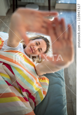 Portrait middle aged woman through palm smiling lying under blanket in bed awaked with good mood. 119947327
