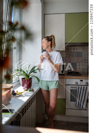 Joyful smiling woman with morning coffee, observing outside from domestic kitchen. Slow morning Joyful smiling woman with morning coffee, observing outside from domestic kitchen. Slow morning 119947339