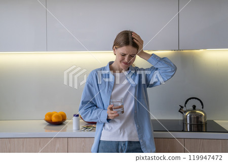 Depressed woman drinks water after taking pills for headache, trying to cope with pain in kitchen 119947472