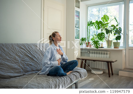 Meditation at home of middle age woman sitting in lotus pose deep breathing touching chest and belly Meditation at home of middle age woman sitting in lotus pose deep breathing touching chest and belly 119947579