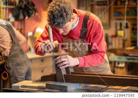 Focused carpenter immersed in work use chisel to smooth bed legs, makes bedframe in workshop 119947595