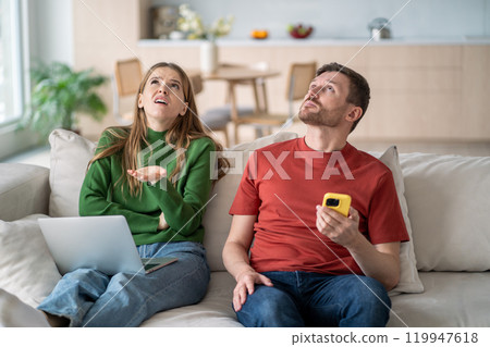 Shocked couple sits on couch with laptop and smartphone, disturbed by from neighbor renovation noise Shocked couple sits on couch with laptop and smartphone, disturbed by from neighbor renovation noise 119947618