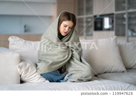 Depressed woman sits on couch wrapped in blanket, feels loneliness, sadness, emotional fatigue, pms 119947628