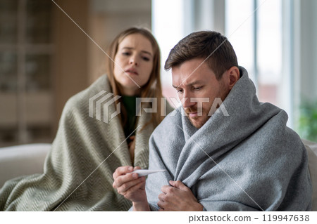 Sick shivering couple of man and woman wrapped in blanket. Male measure fever on couch, battling flu 119947638