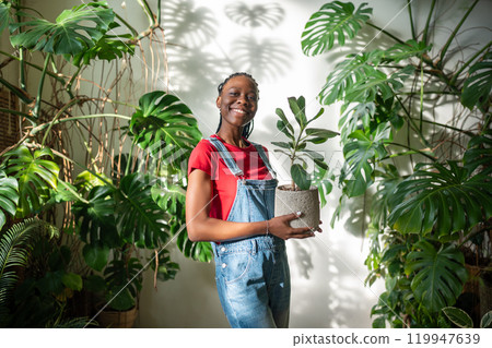 Smiling African American woman holds potted ficus, surrounded by lush flowers, plants in home garden 119947639