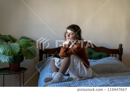 Sad, melancholic woman sits alone on bed, look out window. Feel of loneliness, autumnal melancholy Sad, melancholic woman sits alone on bed, look out window. Feel of loneliness, autumnal melancholy 119947667
