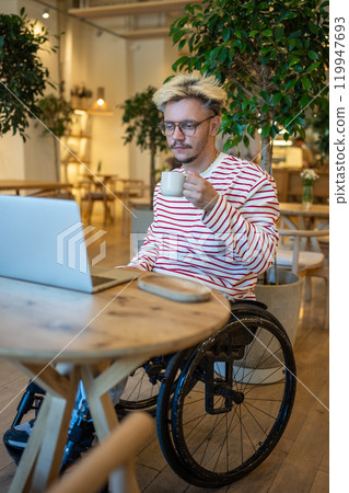 Focused guy freelancer with disabilities in wheelchair working online using laptop sits in cozy cafe 119947693