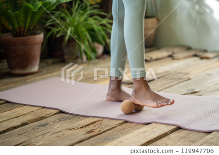 African woman stands on gym mat using massage ball to roll out tension feet spend time for self-care African woman stands on gym mat using massage ball to roll out tension feet spend time for self-care 119947706