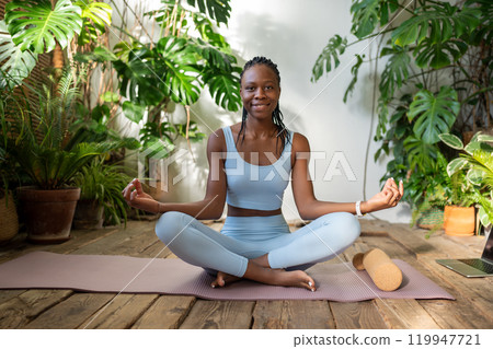 Pleased african american young woman sitting in lotus pose looking at camera in urban jungle place. 119947721