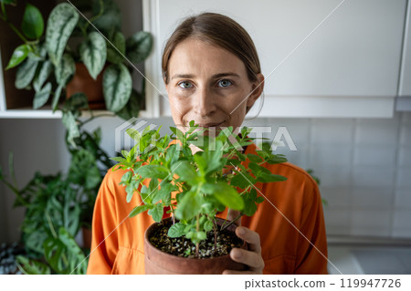 Smiling woman holding flower pot with fresh green mint grown at home, looking at camera. Hobby. Smiling woman holding flower pot with fresh green mint grown at home, looking at camera. Hobby. 119947726