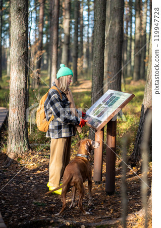 Tourist woman with dog studying information board about wildlife, plants in nature park. Eco tourism Tourist woman with dog studying information board about wildlife, plants in nature park. Eco tourism 119947728