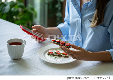 Woman has breakfast with sandwich with cheese and tea, surfing Internet on smartphone in morning Woman has breakfast with sandwich with cheese and tea, surfing Internet on smartphone in morning 119947761